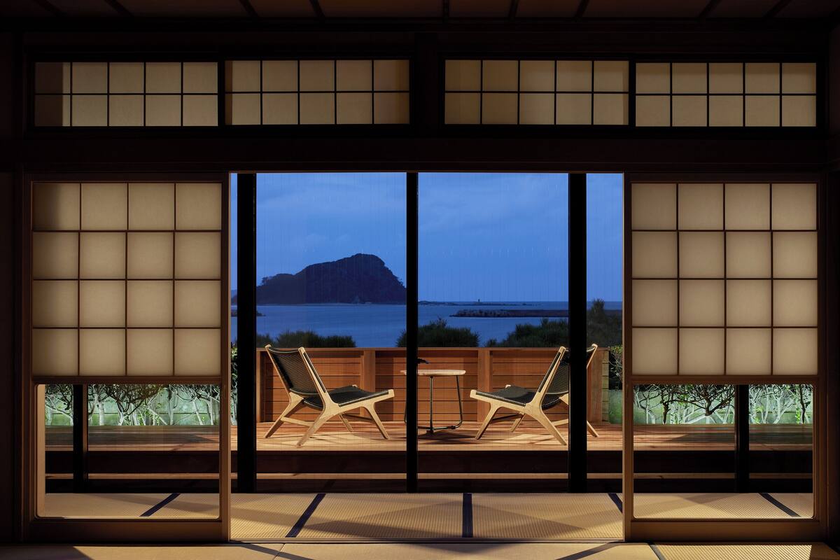 A traditional Japanese room with tatami flooring, a low table, and two chairs facing a large window overlooking a body of water and a distant mountain.