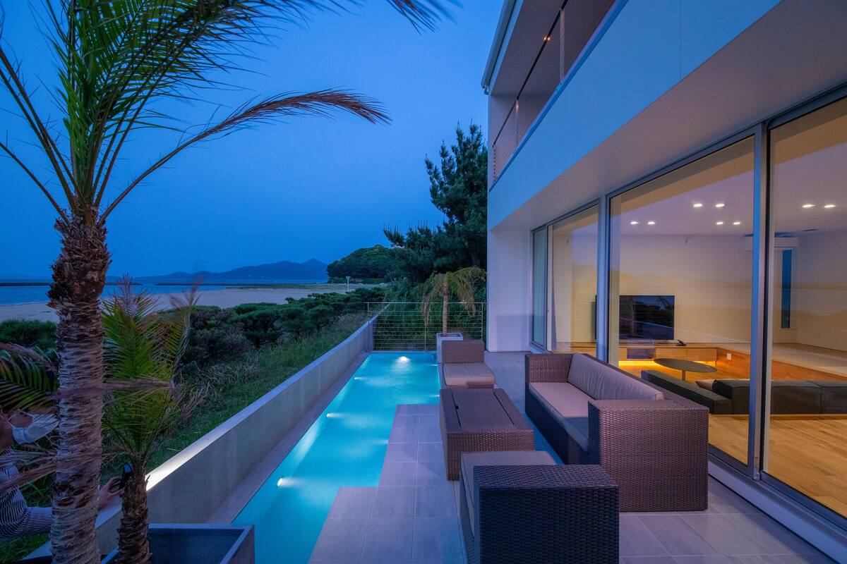 A modern outdoor pool area with seating, a view of the sea, and a large glass wall.