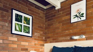 A neatly made bed with white linens and blue accents, a brick wall, and two framed pictures above it. - Red Chief Motel (Gunnedah)