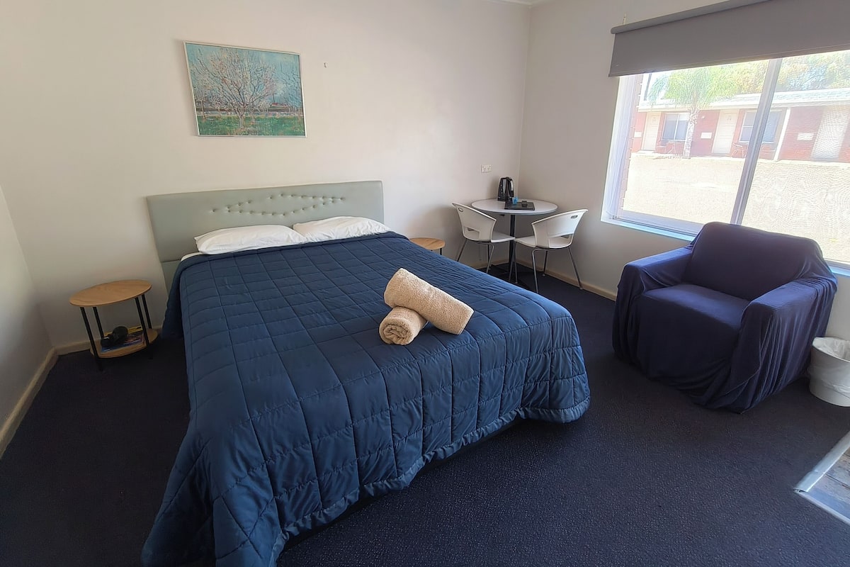 A hotel room with a bed, a small table with chairs, a window, and a painting on the wall.