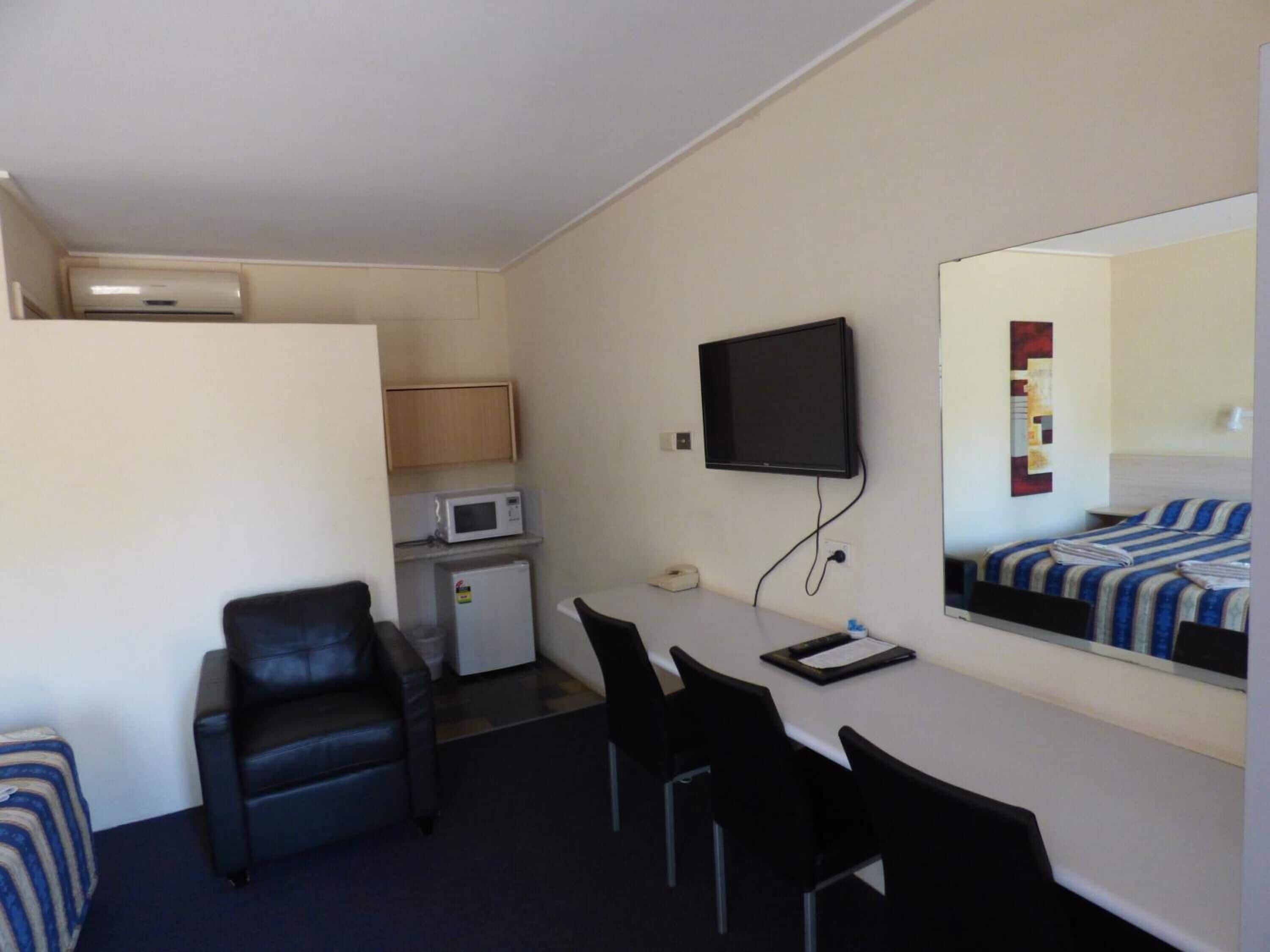 A compact hotel room with a kitchenette, a flat-screen TV mounted on the wall, a bed with striped bedding, and a seating area with chairs.