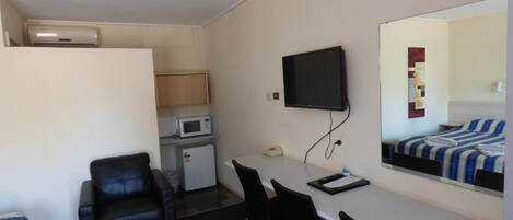 A compact hotel room with a kitchenette, a flat-screen TV mounted on the wall, a bed with striped bedding, and a seating area with chairs.