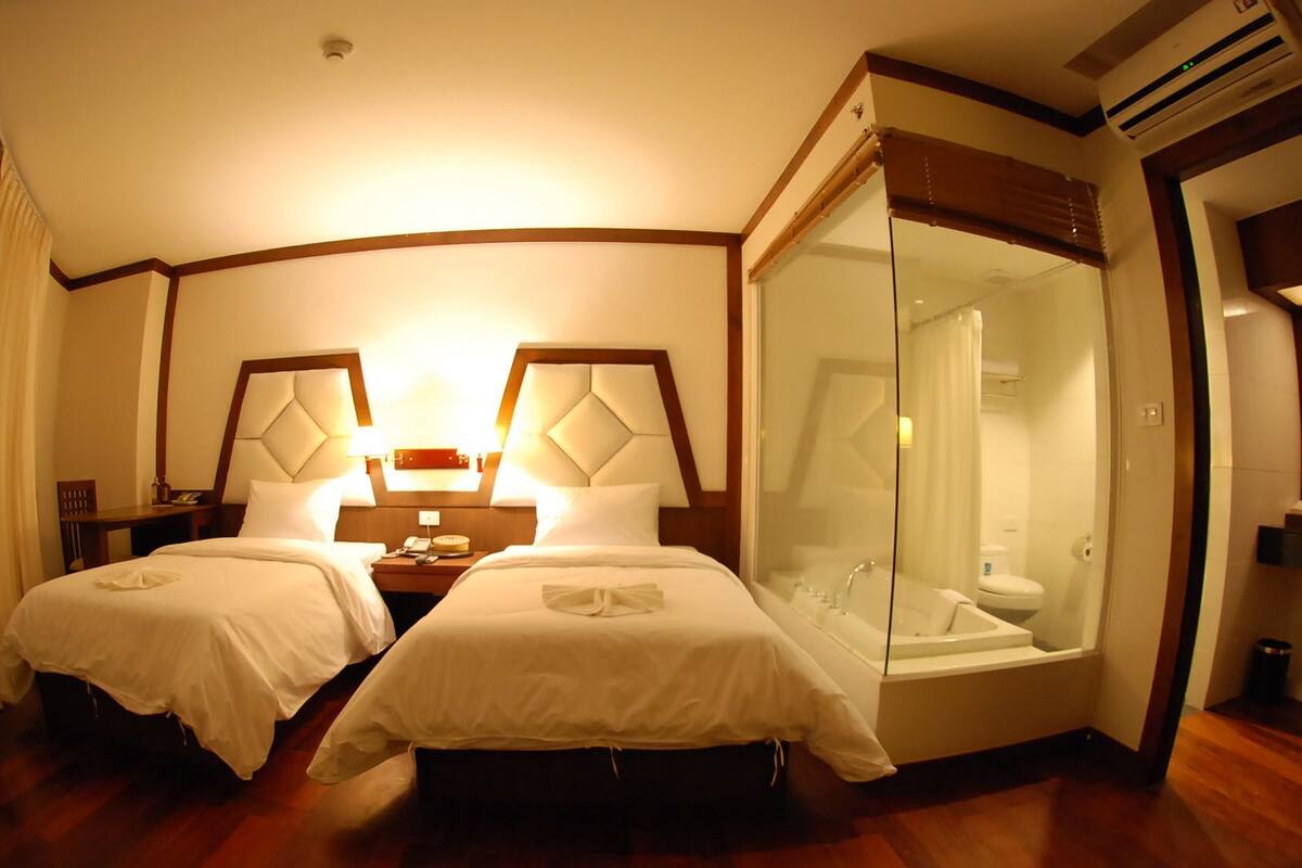 A hotel room with two beds, a glass-enclosed bathroom, and a wooden floor.