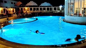 Una piscina con dos personas nadando, rodeada de sillas de descanso y un edificio con un ventanal grande.
