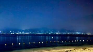 Een nachtelijk kustlandschap met een stadshemelbeeld op de achtergrond, een zandstrand op de voorgrond en een kalme zee die de lichtjes weerspiegelt.