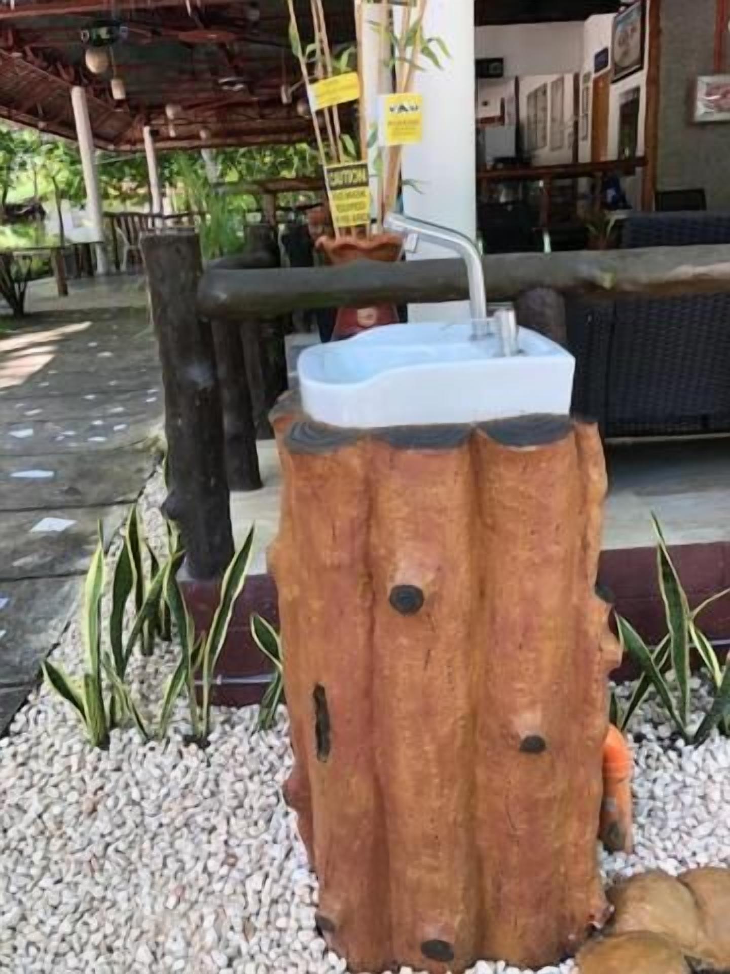 Un lavabo original, en forme de tronc d’arbre, doté d’un robinet et entouré de petits galets blancs et de plantes.