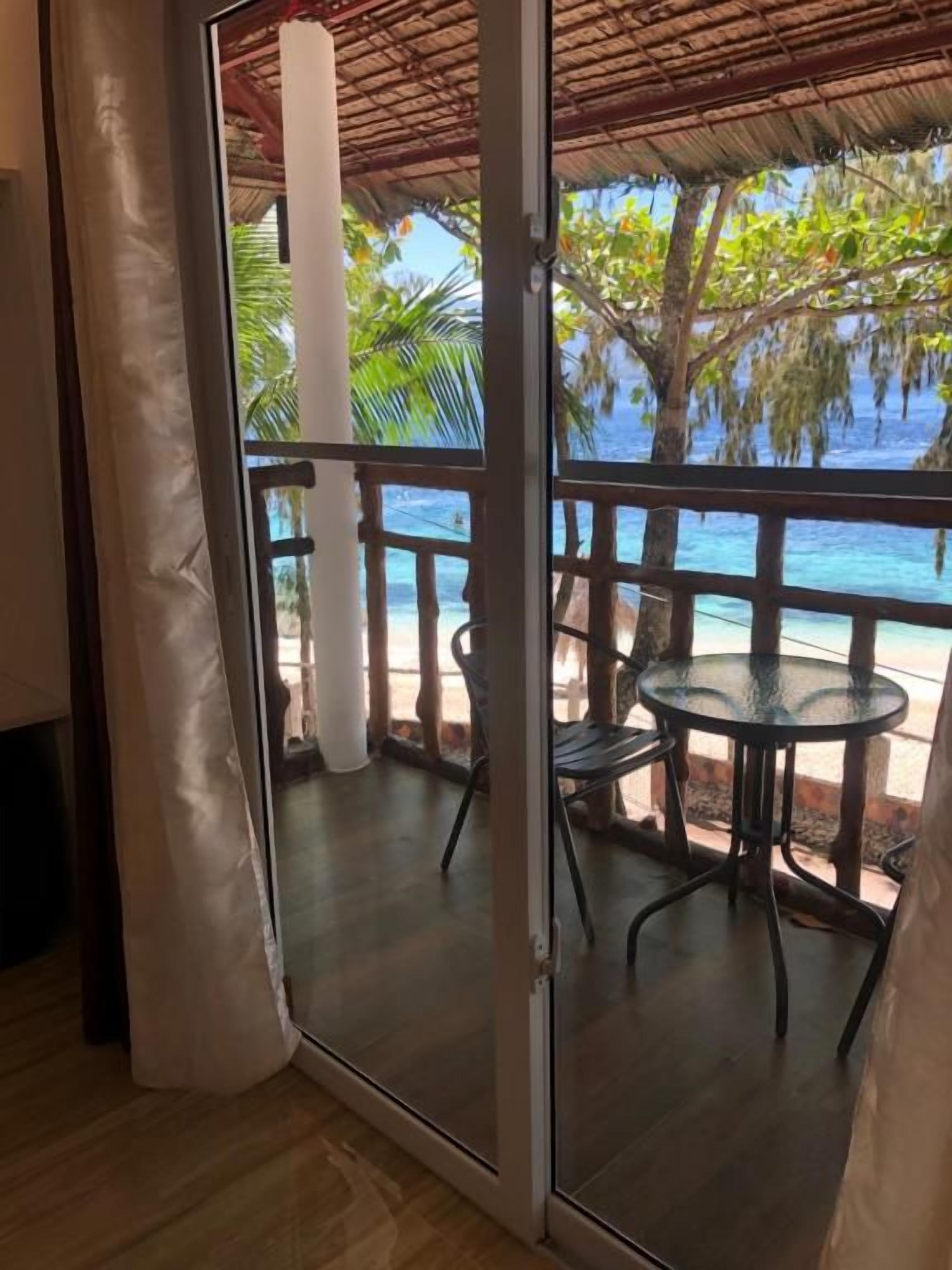 A room with a glass door leading to a balcony with a table and chairs, overlooking a beach and palm trees.