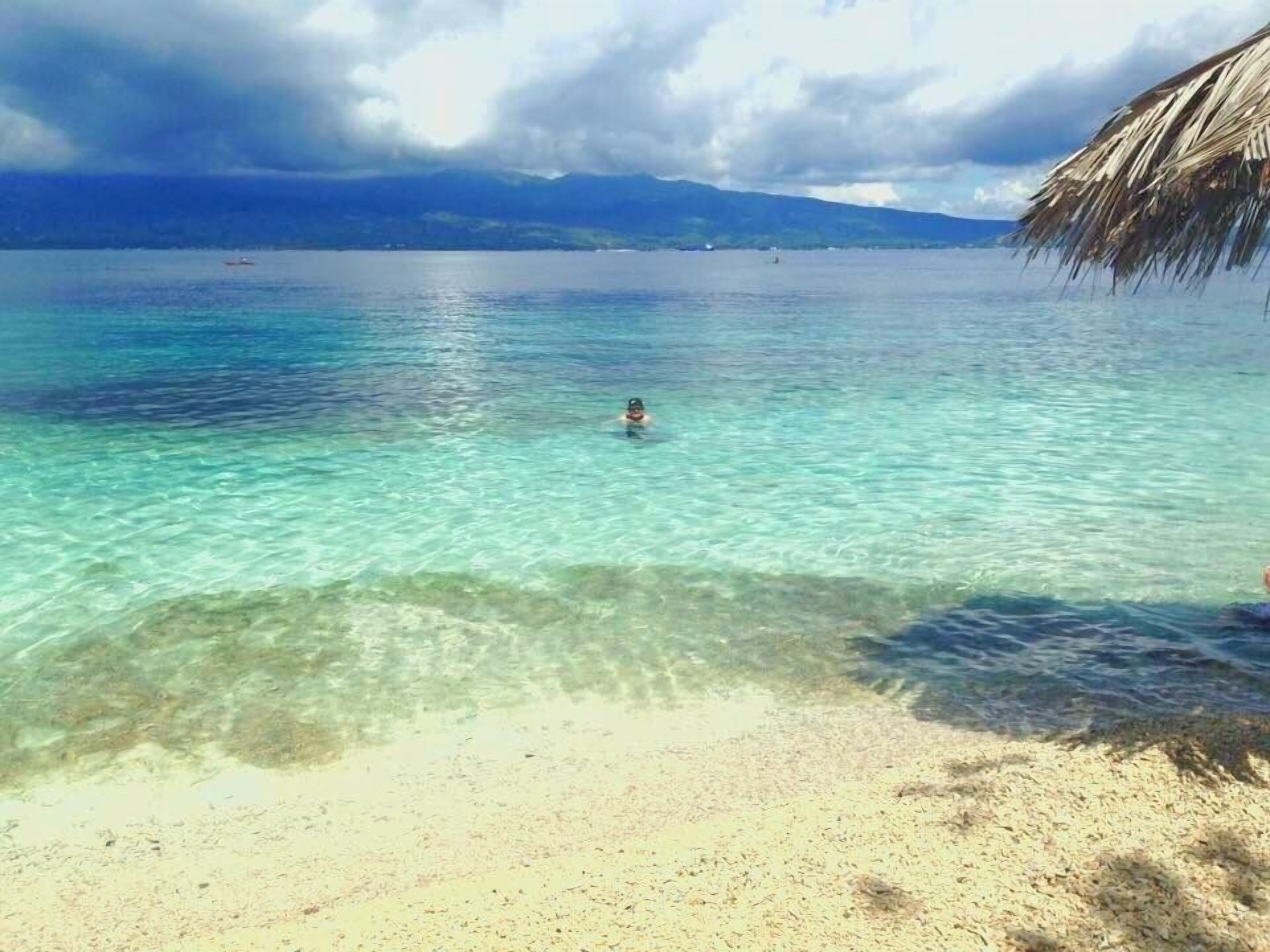 Una playa con agua turquesa cristalina, una persona nadando y una estructura de paja a la derecha.