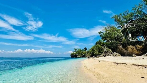 Une plage à l’eau turquoise et limpide, un rivage sablonneux et un bâtiment doté d’un balcon donnant sur la mer.