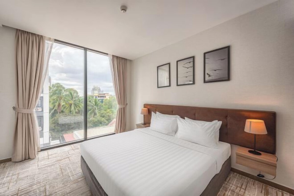 Una habitación de hotel con una cama grande, una mesita de noche con lámpara, una ventana con cortinas y cuadros en la pared.