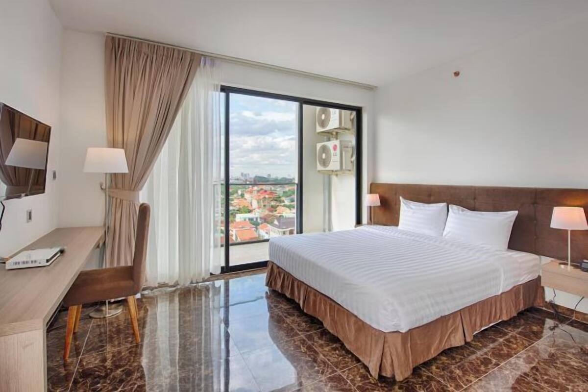 Habitación de hotel con cama, escritorio, silla, ventana grande con cortinas y un equipo de aire acondicionado.