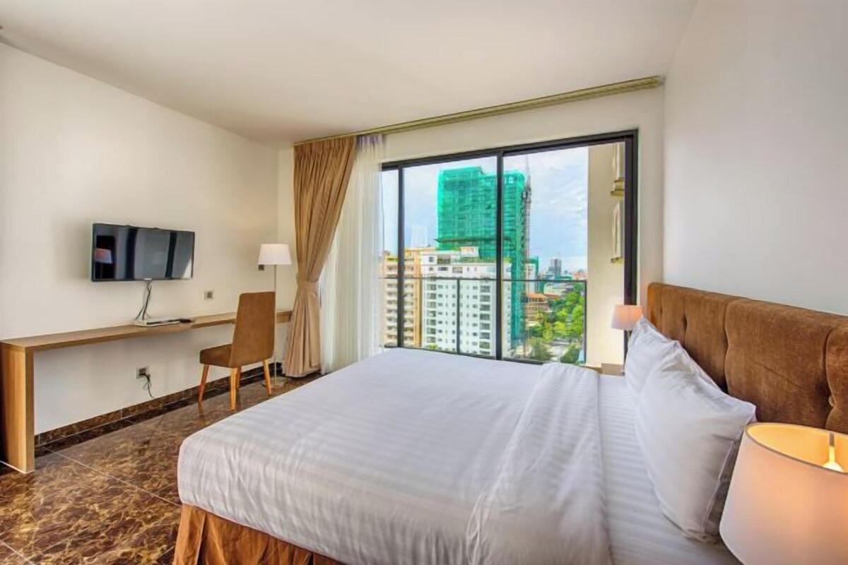 Habitación de hotel con cama, escritorio, silla, televisión y ventana grande con vistas a edificios.