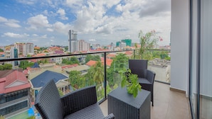 Un balcón con mobiliario de mimbre y una planta en maceta, con vistas a un paisaje urbano con diversos edificios y un cielo despejado.