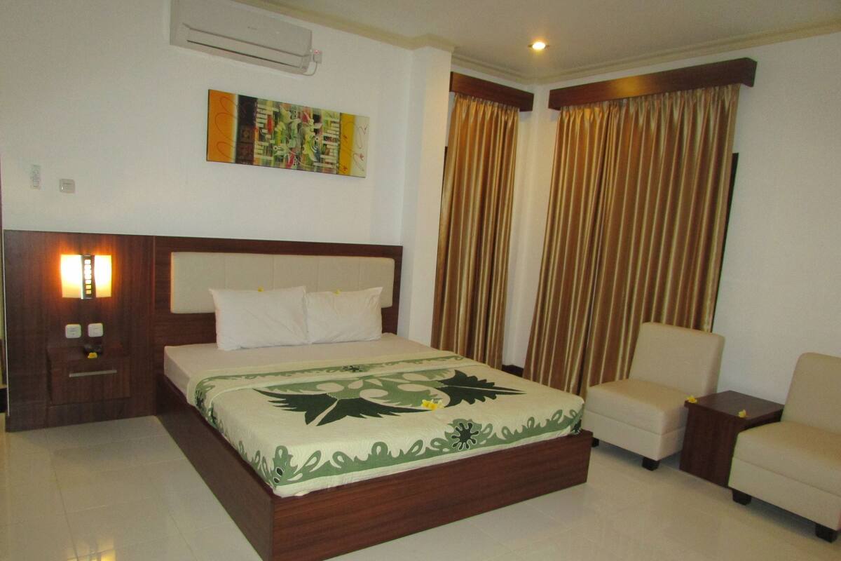 A hotel room with a bed, a chair, a nightstand, a wall-mounted air conditioner, and a painting.