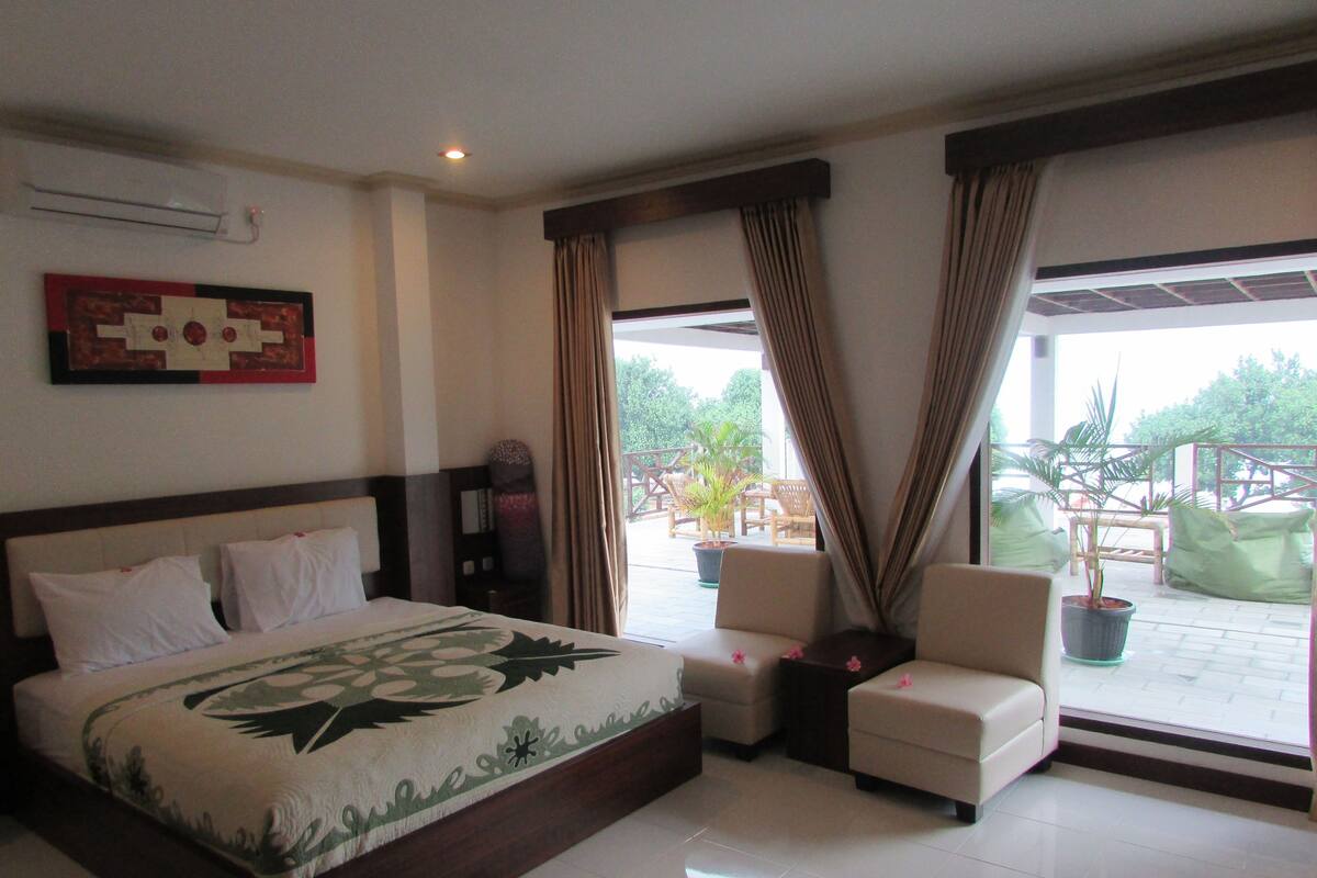 A hotel room with a bed, two chairs, a potted plant, and a view of the outdoors.