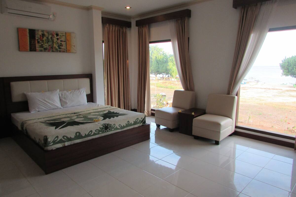 A hotel room with a large bed, two armchairs, a small table, and a view of the beach.