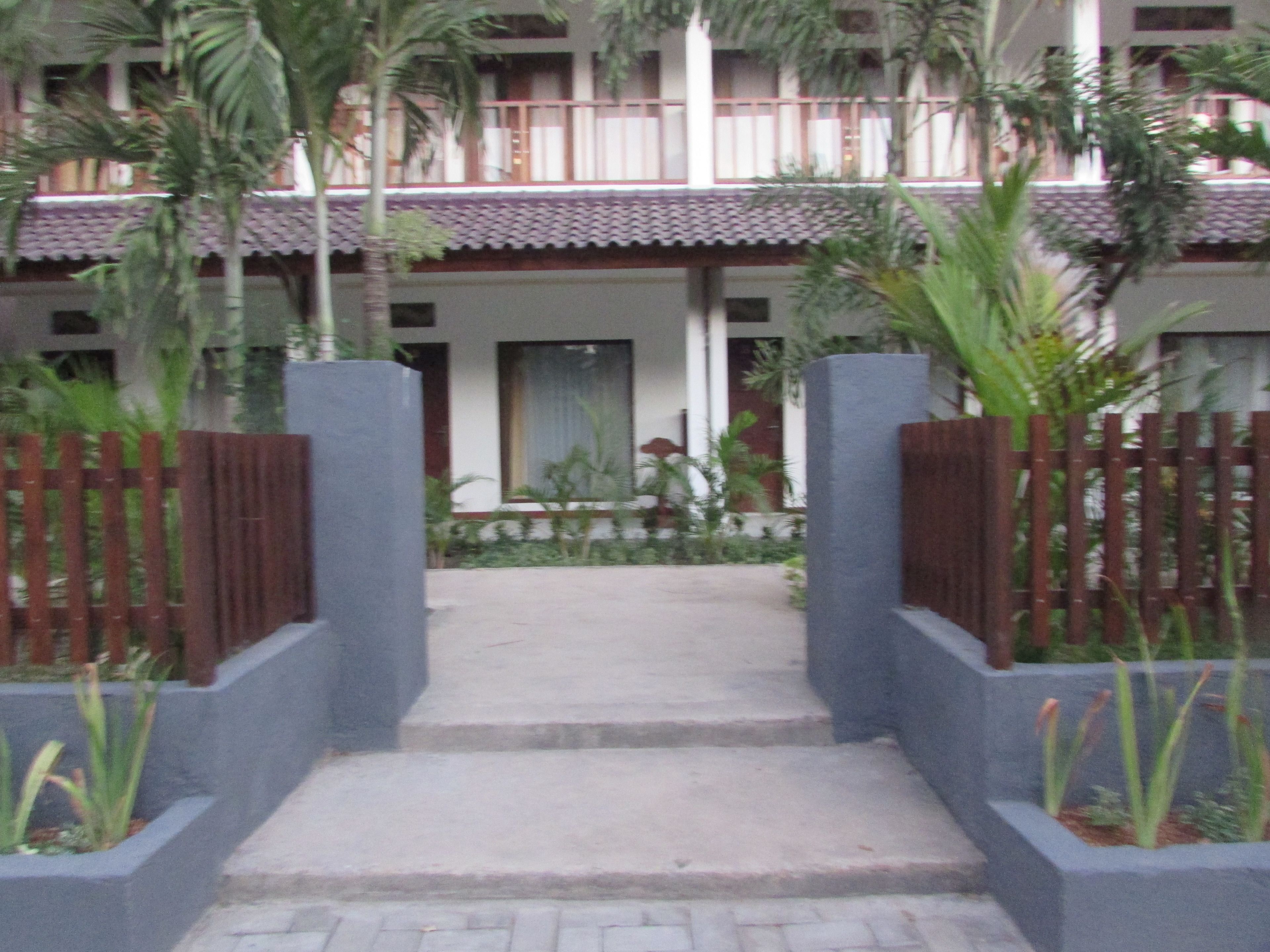 A hotel entrance with a concrete pathway leading to a door, surrounded by a wooden fence and greenery.