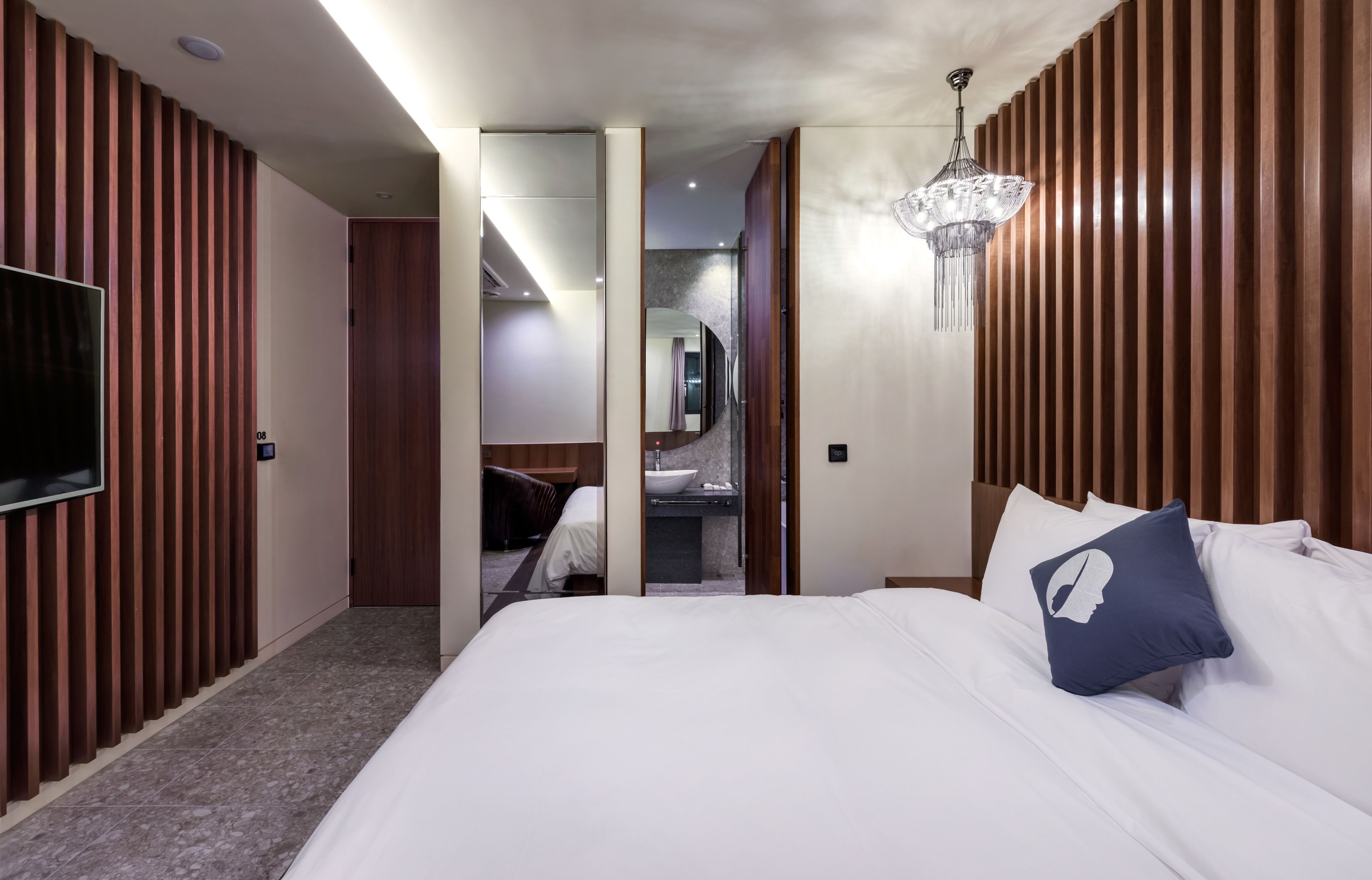 A modern hotel room with a large bed, a TV, a chandelier, and a bathroom visible through an open door.