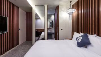 A modern hotel room with a large bed, a TV, a chandelier, and a bathroom visible through an open door.