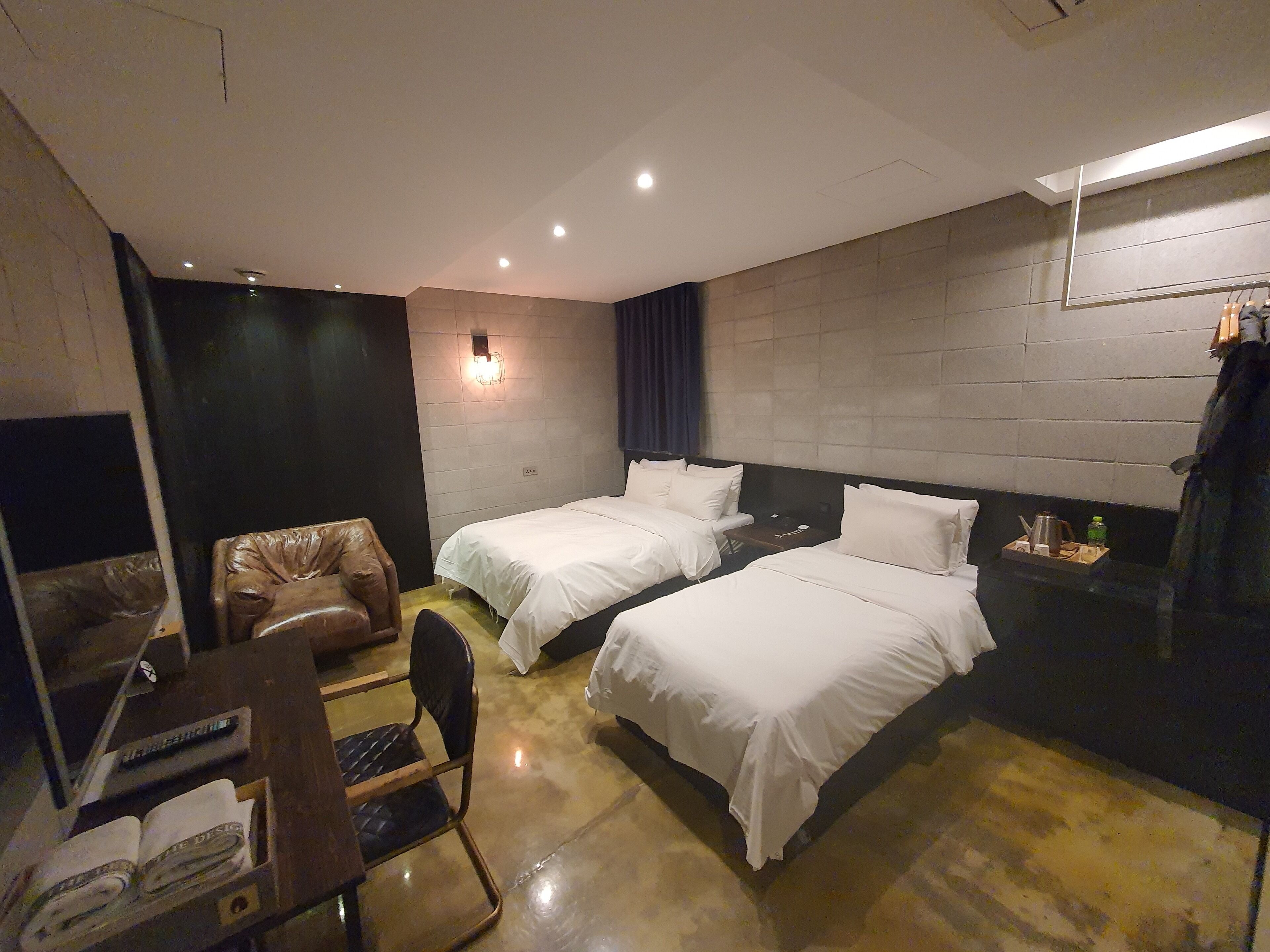 Habitación de hotel con dos camas, un escritorio con silla, una mesita con hervidor y estante en la pared.
