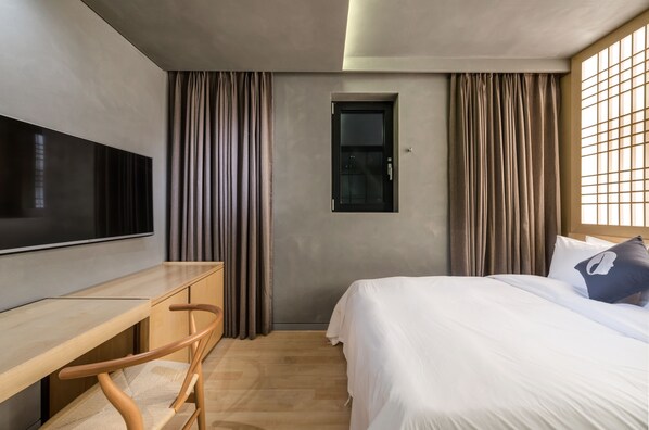 Habitación de hotel moderna con televisión de pantalla plana, escritorio de madera, una butaca cómoda, cama con ropa blanca y ventana con cortinas.