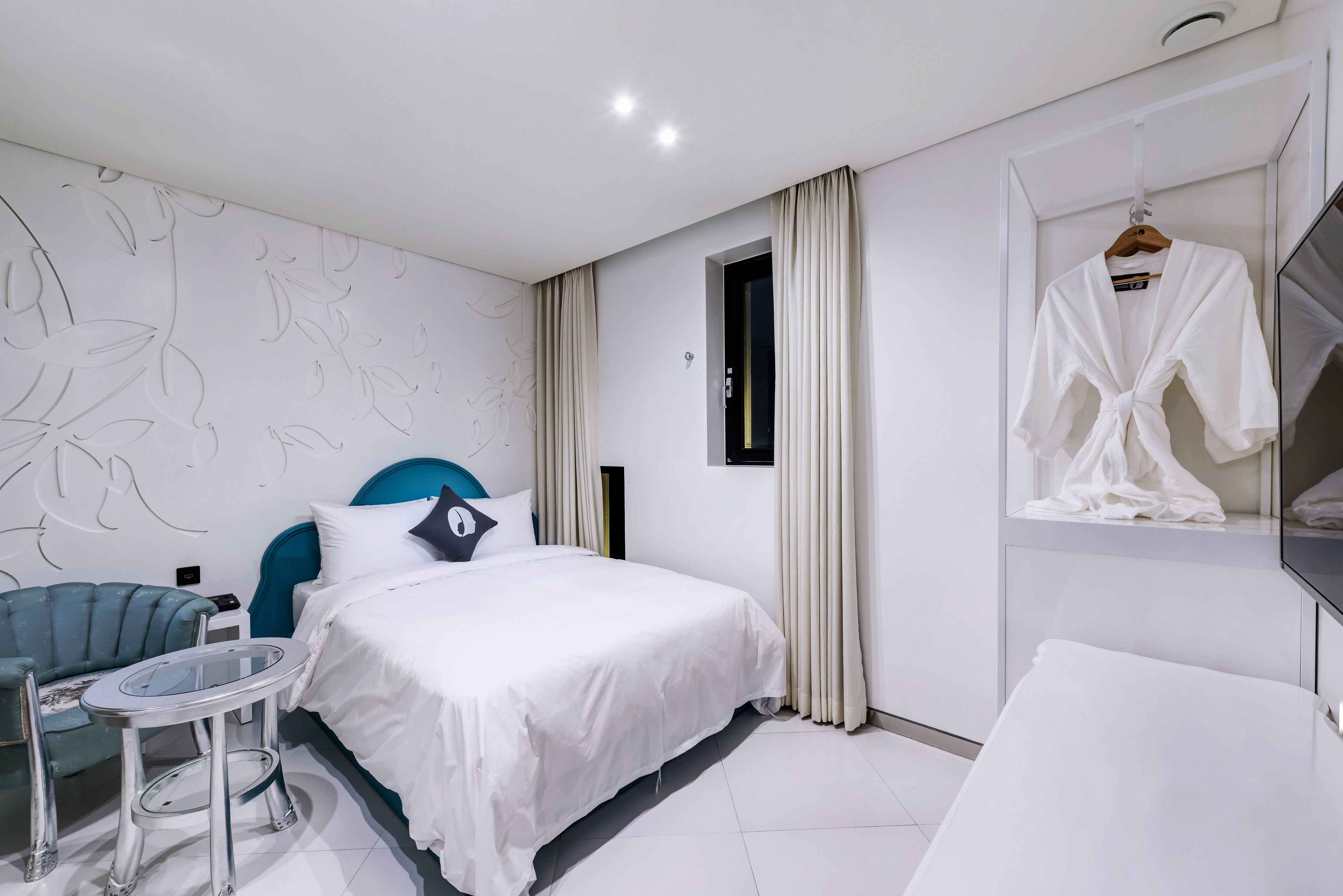 Habitación de hotel moderna con una cama, una mesa redonda, un sillón azul y una bata colgada en un perchero.
