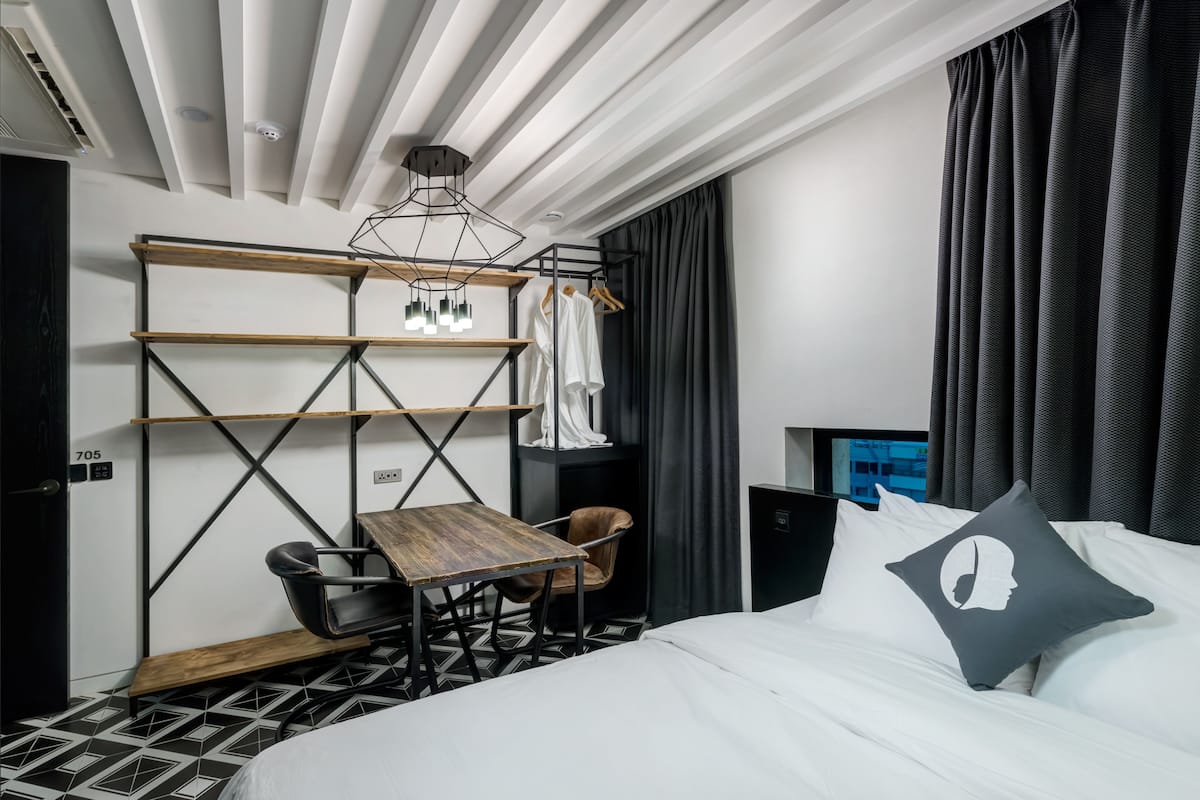 A modern hotel room with a bed, a desk, a chair, a wardrobe, and a hanging light fixture.