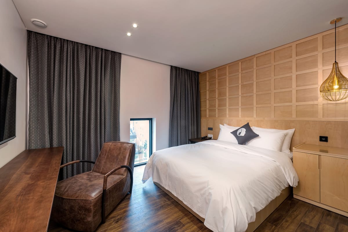 A modern hotel room with a large bed, a wooden headboard, a desk, and a chair.