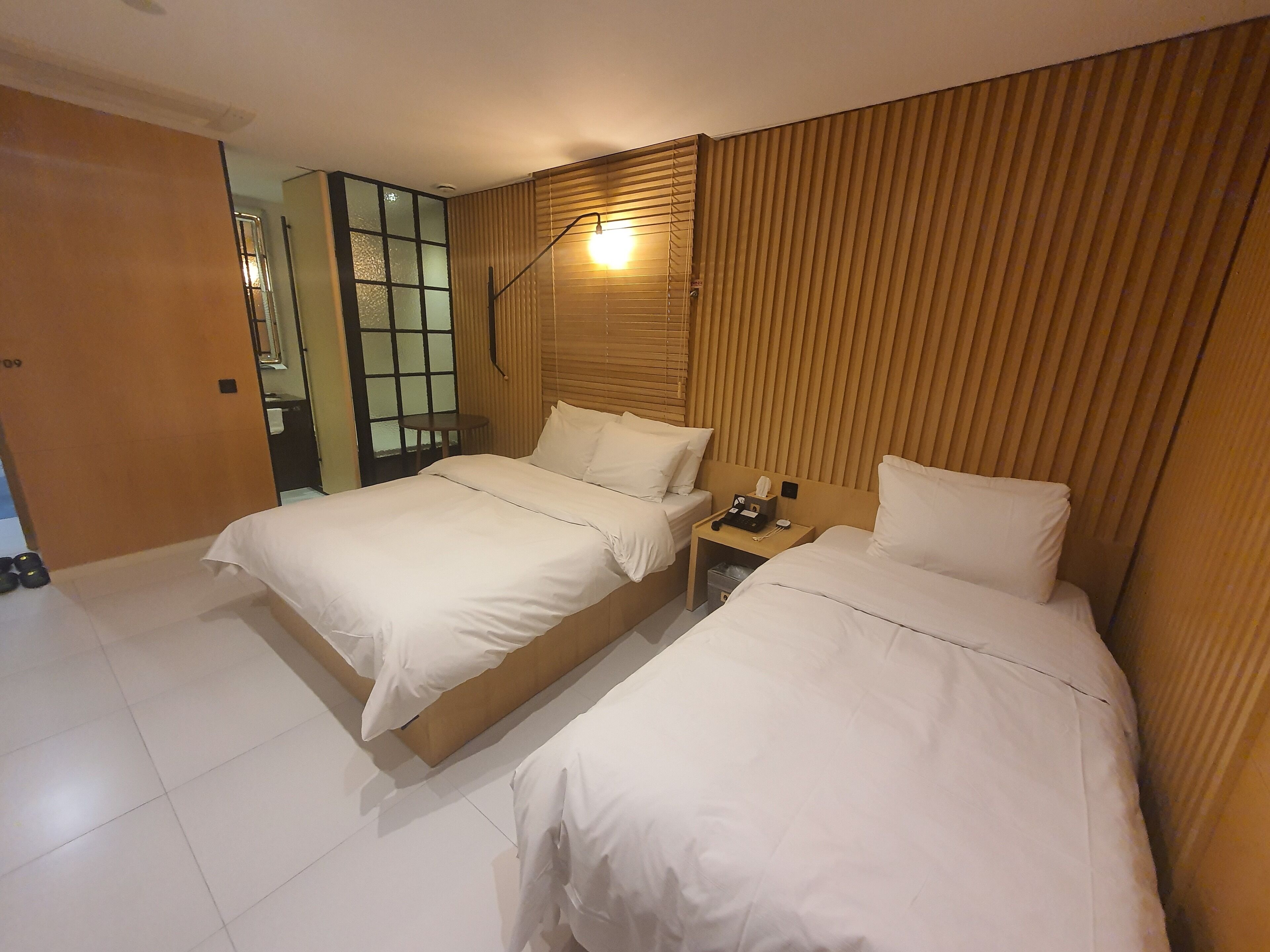 Habitación de hotel con dos camas, un cabecero de madera, una mesita de noche con lámpara y una pequeña mesa.