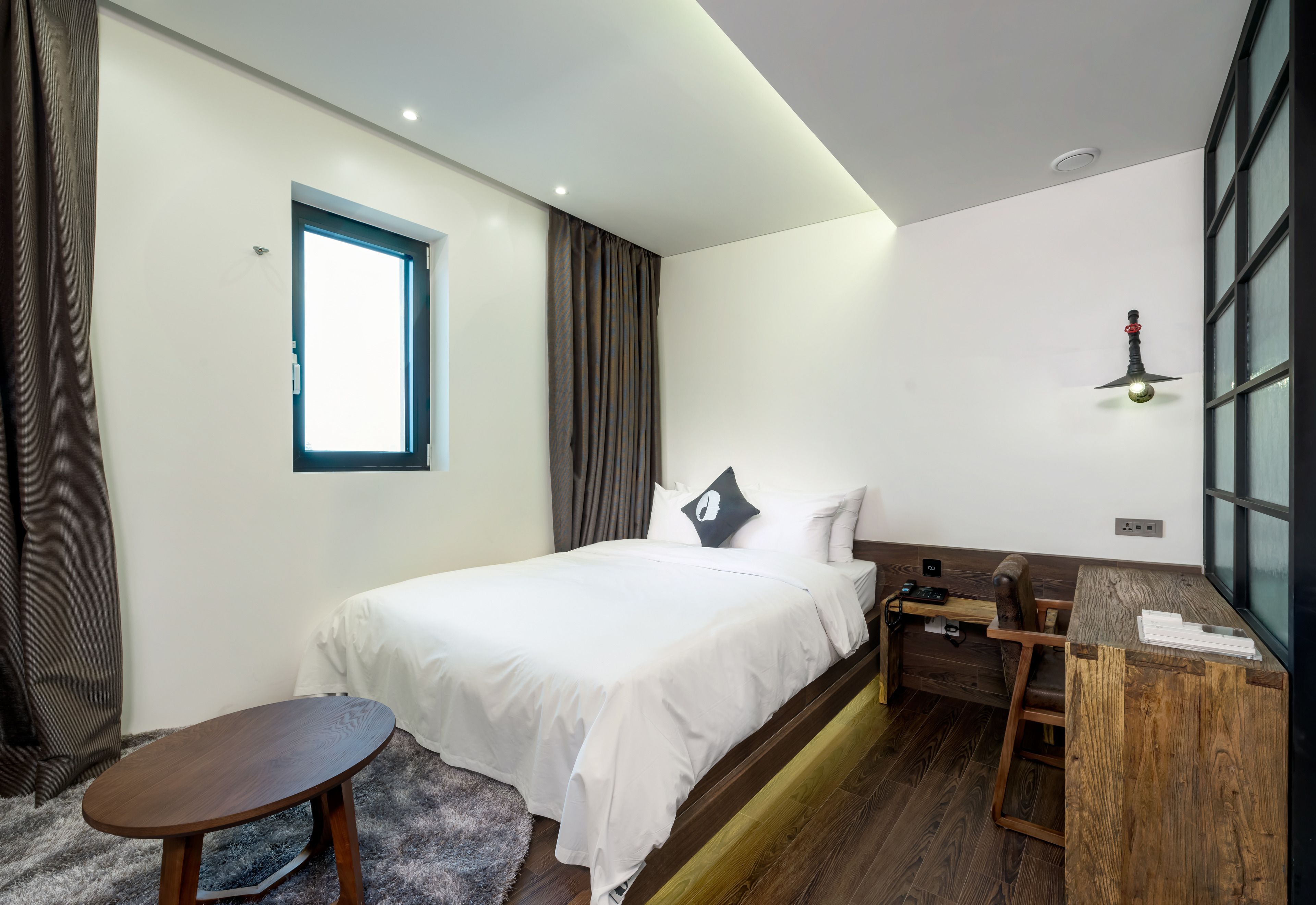 A modern hotel room with a bed, a small wooden desk, a window, and a ceiling light fixture.
