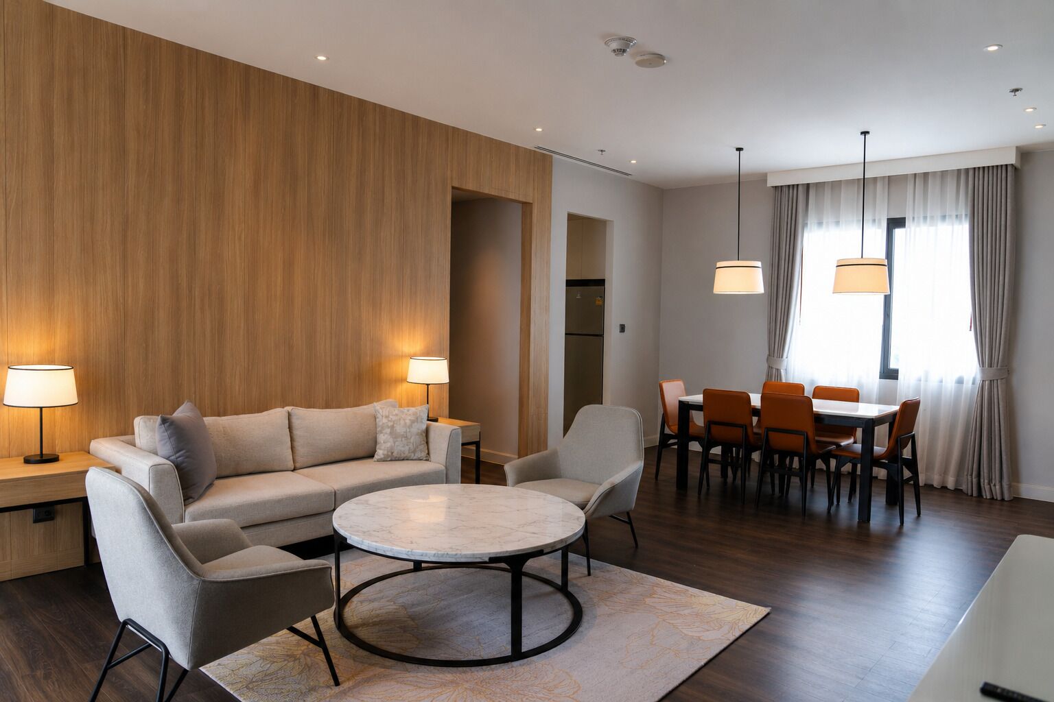Une chambre d’hôtel moderne équipée d’un canapé, de fauteuils, d’une table ronde, d’un coin repas avec des chaises et de luminaires suspendus.
