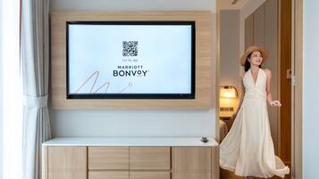 Habitación de hotel con televisión de pantalla plana en la pared, una mesita de madera y una persona con vestido blanco caminando hacia la puerta.