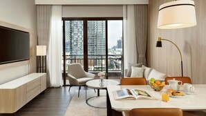 Una habitación de hotel moderna con un ventanal grande, un televisor de pantalla plana, una zona de comedor con una mesa preparada para el desayuno y un espacio de estar acogedor con un sofá y sillones.