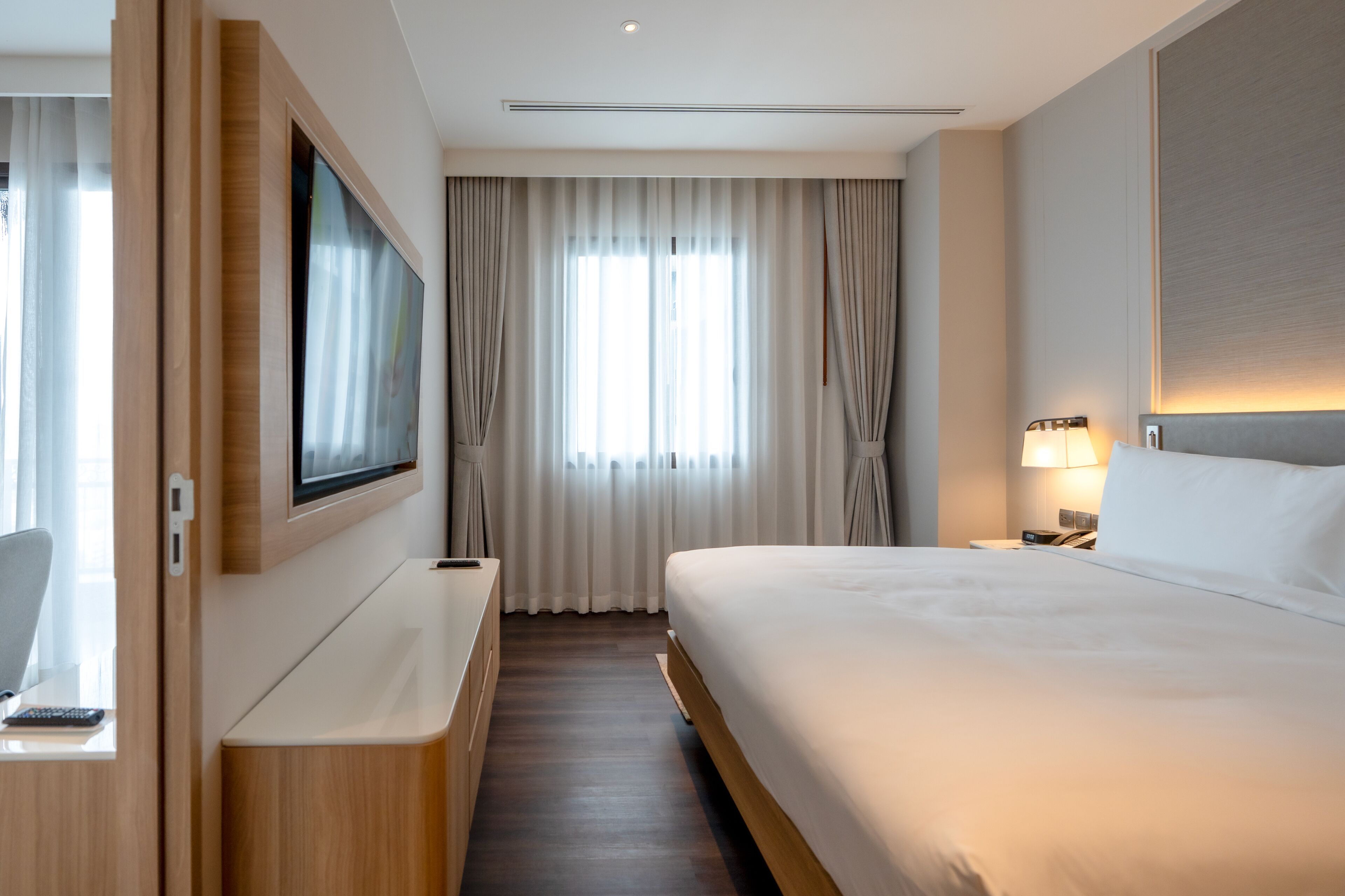 A modern hotel room with a large bed, a flat-screen TV mounted on the wall, a bedside table with a lamp, and a view of the outdoors through sheer curtains.