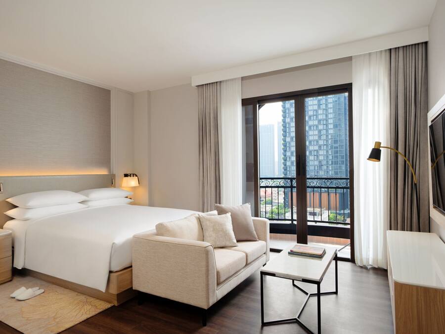 A hotel room with a large bed, a sofa, a small table, a TV, and a balcony with a city view.
