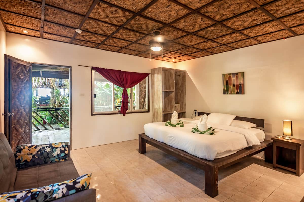 Une chambre avec un lit en bois, un canapé, une fenêtre avec des rideaux, un plafond orné de motifs en bois et un tableau accroché au mur.