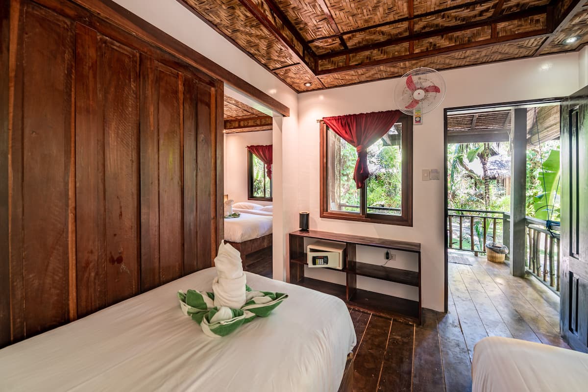 Une chambre avec deux lits, un plafond en bois, un ventilateur, une fenêtre donnant sur un paysage verdoyant et un balcon.