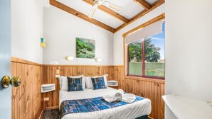 A bedroom with a wooden ceiling, a bed with a blue and white bedspread, a window with a view of greenery, and a white bathtub. - Discovery Parks - Grampians (Halls Gap)