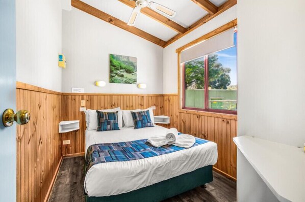 A bedroom with a wooden ceiling, a bed with a blue and white bedspread, a window with a view of greenery, and a white bathtub. - Breeze Holiday Parks - Grampians (Halls Gap)