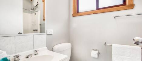 Un baño con lavamanos, inodoro y espejo blancos. Hay una ventana sobre el inodoro y un perchero en la pared.