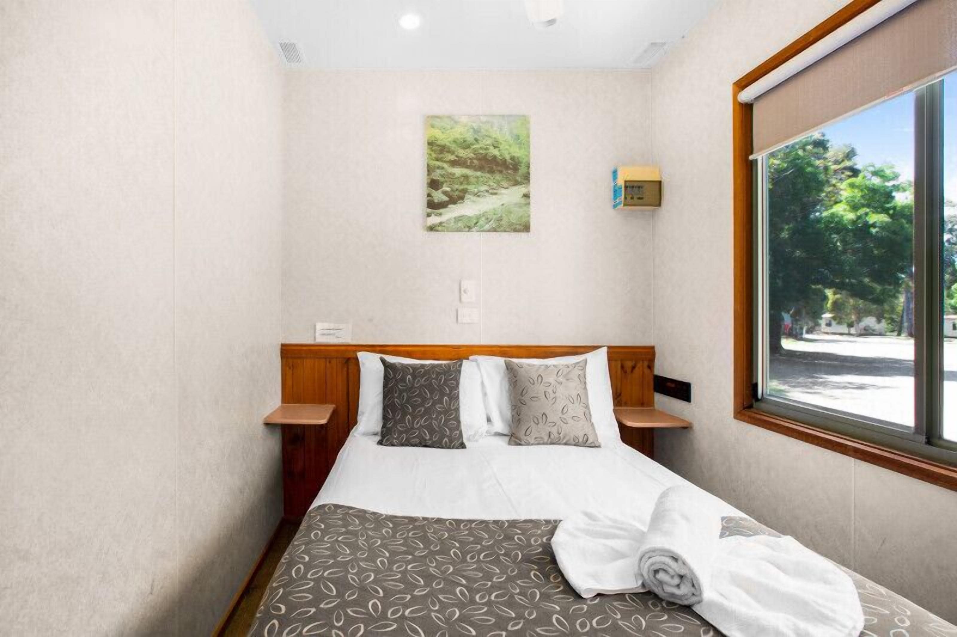 A neatly made bed with patterned bedding, a wall-mounted picture, and a window with a view of trees.