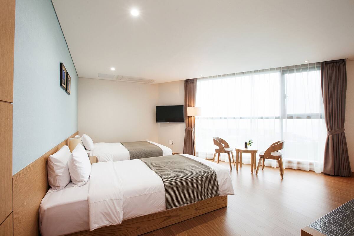 A hotel room with two beds, a TV, a small table with chairs, and a large window with curtains.