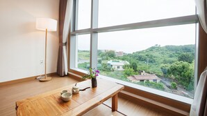 A room with a wooden table, a vase with flowers, a lamp, and a large window with a view of greenery and buildings. - You Us Hotel (Seogwipo)