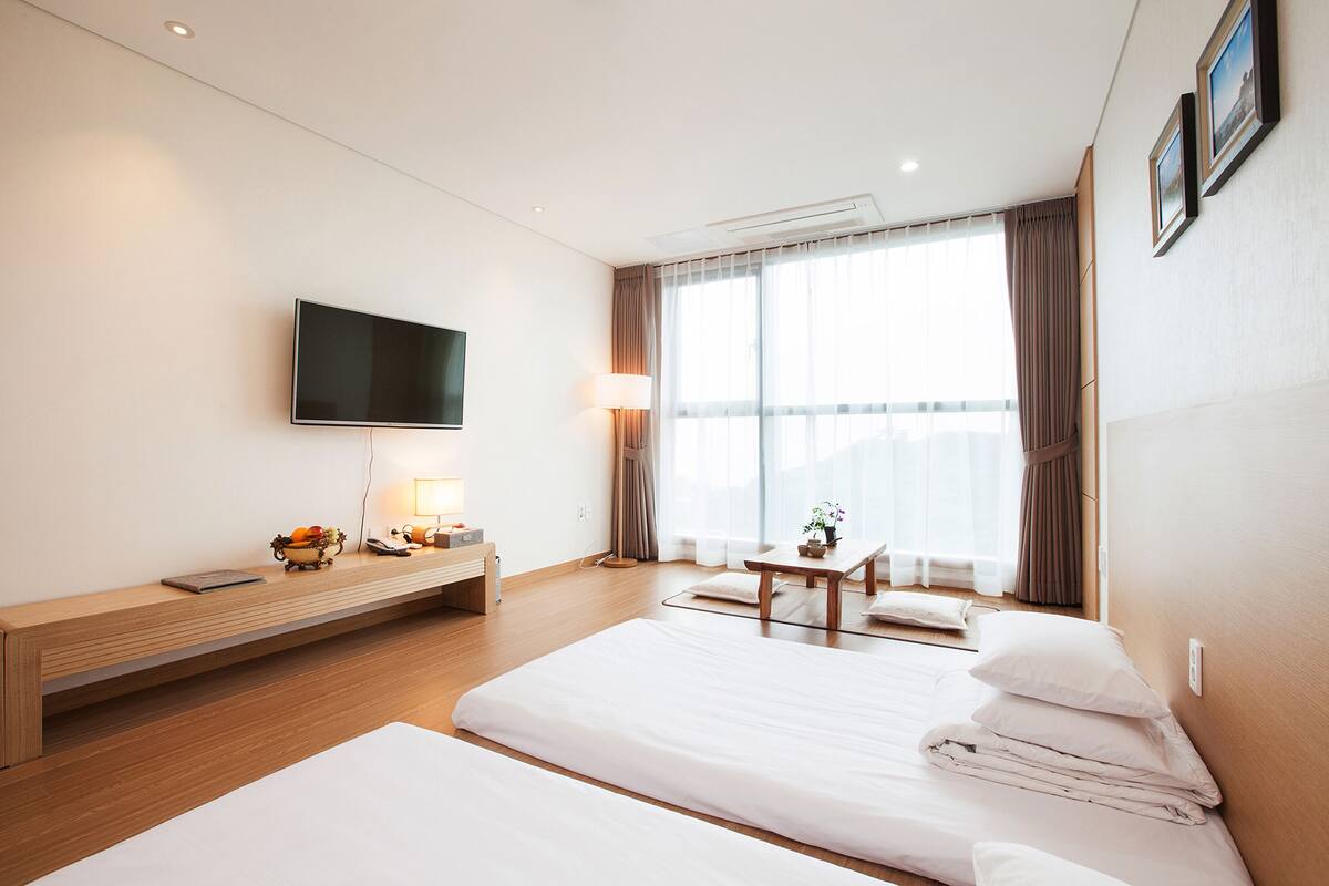 A hotel room with a large bed, a smaller bed, a TV mounted on the wall, a desk with a lamp, and a window with curtains.