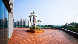 A rooftop terrace with wooden flooring, a central planter with palm trees, and a cityscape view. - You Us Hotel (Seogwipo)