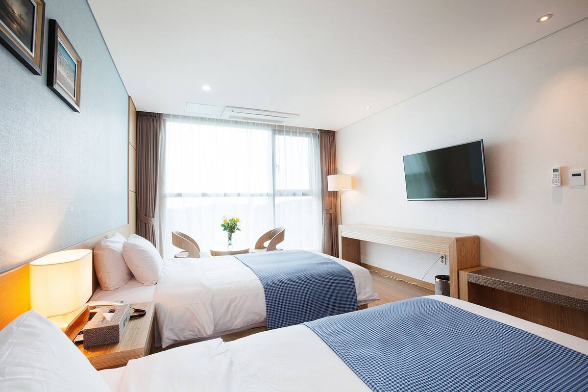 A hotel room with two beds, a desk with a lamp, a TV on the wall, and a window with curtains.