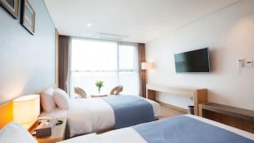 Ein Hotelzimmer mit zwei Betten, einem Schreibtisch mit Lampe, einem Wandfernseher und einem fenster mit Vorhängen.