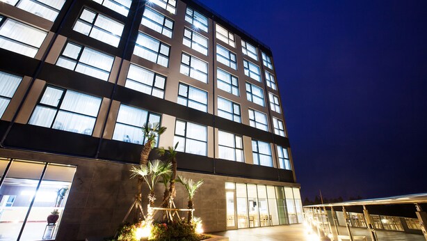 A modern building with multiple floors, large windows, and a well-lit entrance area with a small garden. - You Us Hotel (Seogwipo)