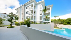 A modern multi-story building with a swimming pool, surrounded by greenery and palm trees.