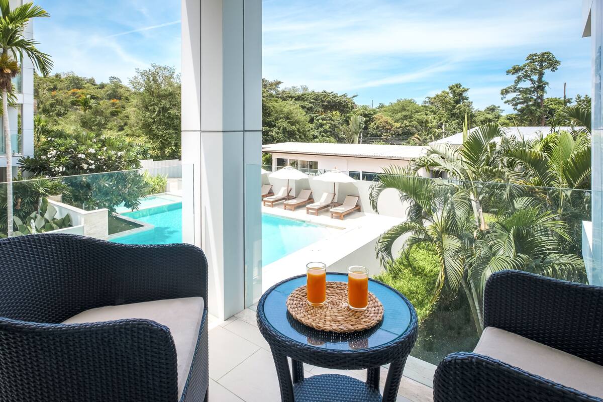 A balcony with wicker furniture, a glass table with two drinks, and a view of a pool and lush greenery.